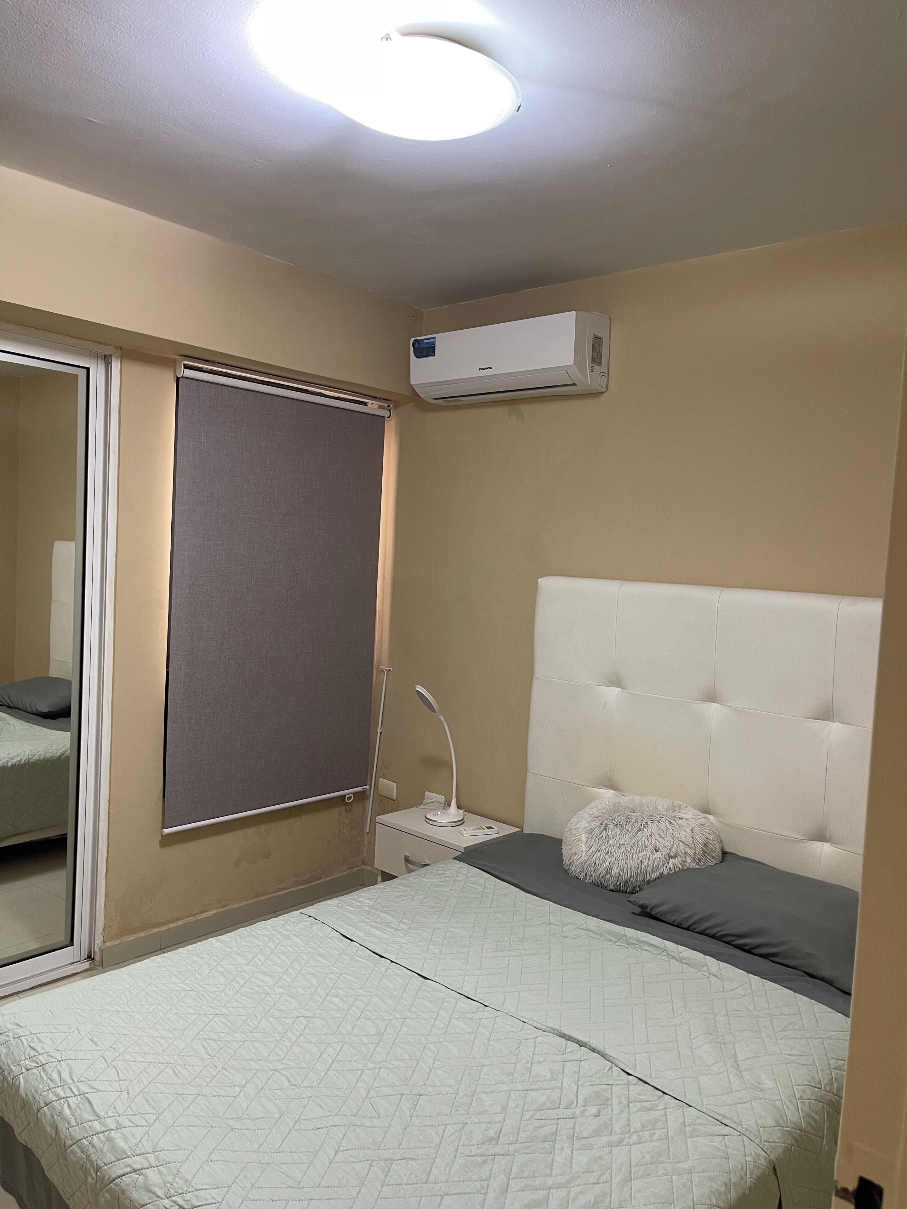 A bedroom with a bed, a headboard, a bedside table with a lamp, a window with a blind, and an air conditioning unit on the wall.