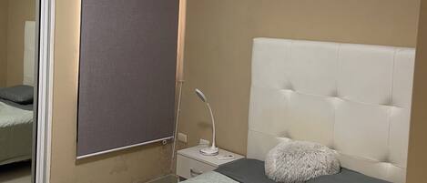 A bedroom with a bed, a headboard, a bedside table with a lamp, a window with a blind, and an air conditioning unit on the wall.