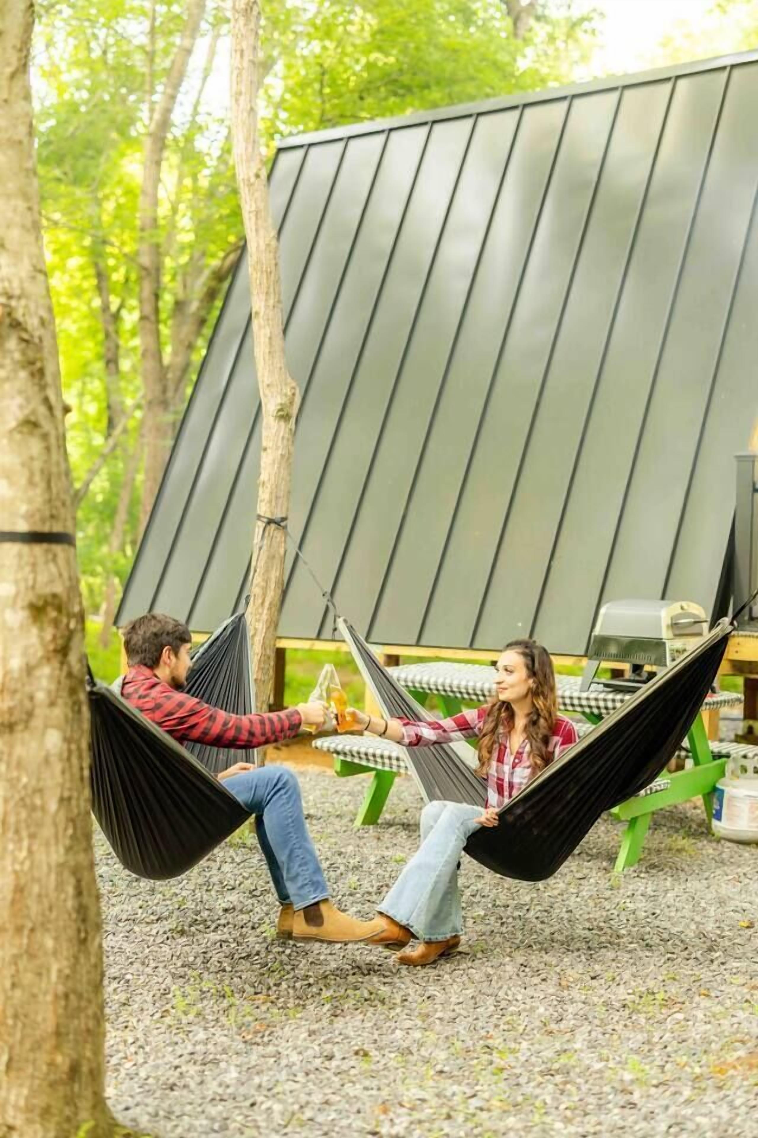 Two people relaxing in hammocks under a modern, sloped-roof structure surrounded by trees.