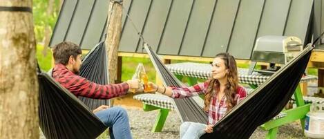 Two people relaxing in hammocks under a modern, sloped-roof structure surrounded by trees.