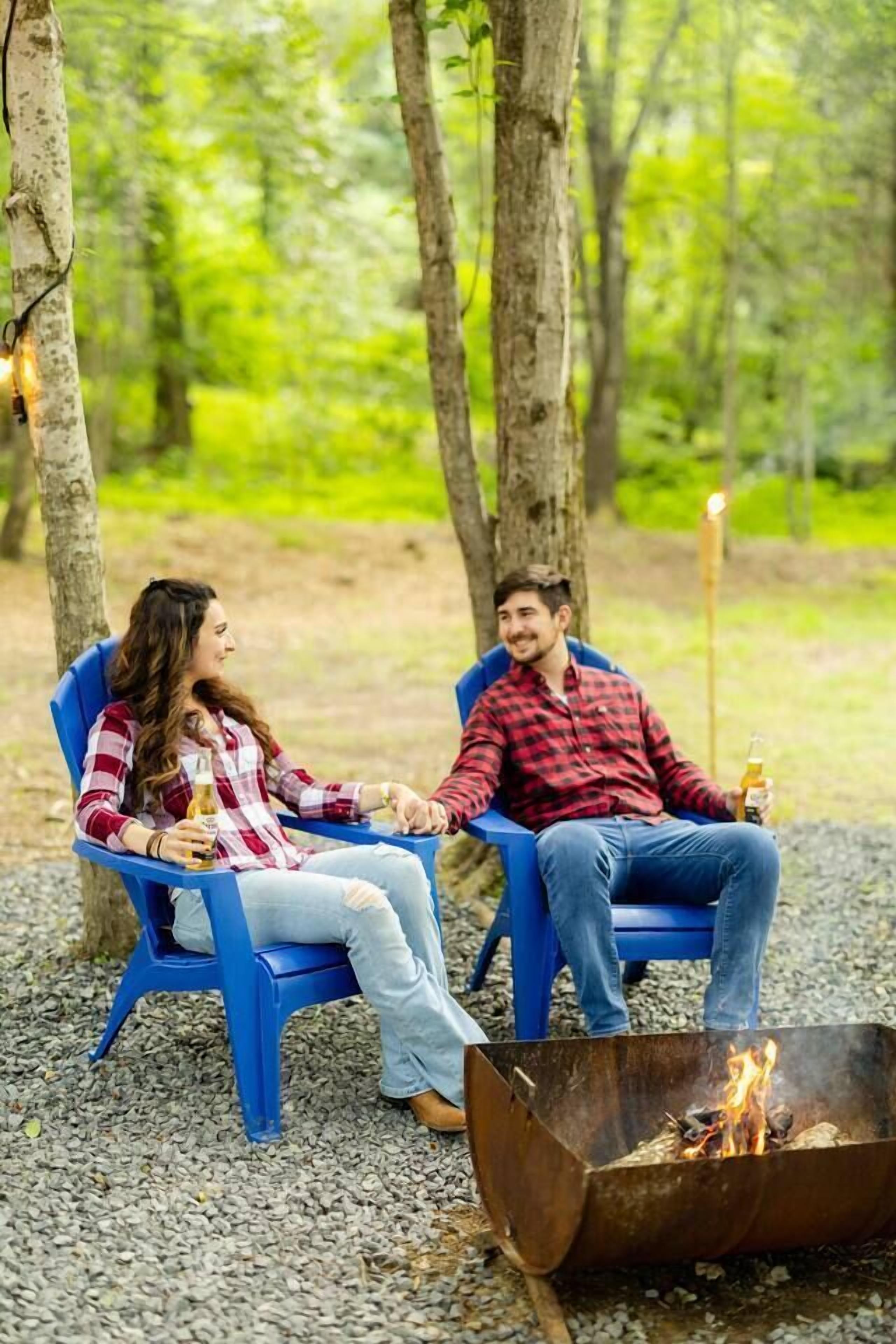 Deux personnes sont assises sur des chaises bleues près d’un feu de camp, dans un cadre forestier.