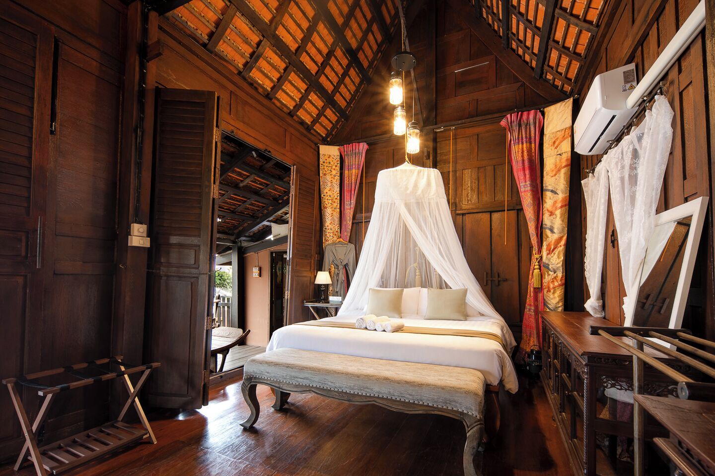 A traditional wooden room with a bed under a canopy, a wooden bench, and a hanging air conditioner.