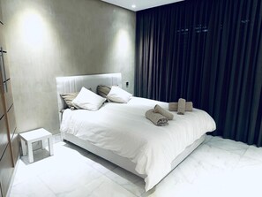 Et moderne hotelværelse med en stor seng, et lille natbord og mørke gardiner.