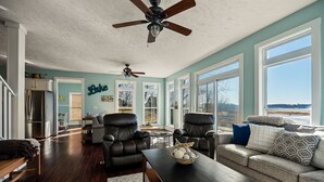 Ein großzügiges Wohnzimmer mit dunklem Holzboden, einem Sofa, einem Couchtisch und zwei Ledersesseln. Im Hintergrund sind zwei Deckenventilatoren, ein Kühlschrank und eine Küchenzeile zu sehen.
