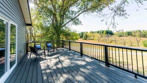 A wooden deck with a black railing, a table with chairs, and a view of trees and open land.