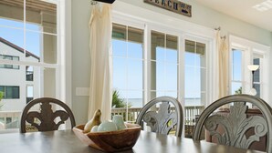A dining table with a bowl of fruit, white curtains, and a view of the ocean through large windows. - Water Views + Pool Access | Driftwood Cottage (Navarre Beach)