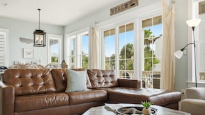 A living room with a brown leather sectional sofa, a coffee table with decorative items, and large windows with curtains. - Water Views + Pool Access | Driftwood Cottage (Navarre Beach)