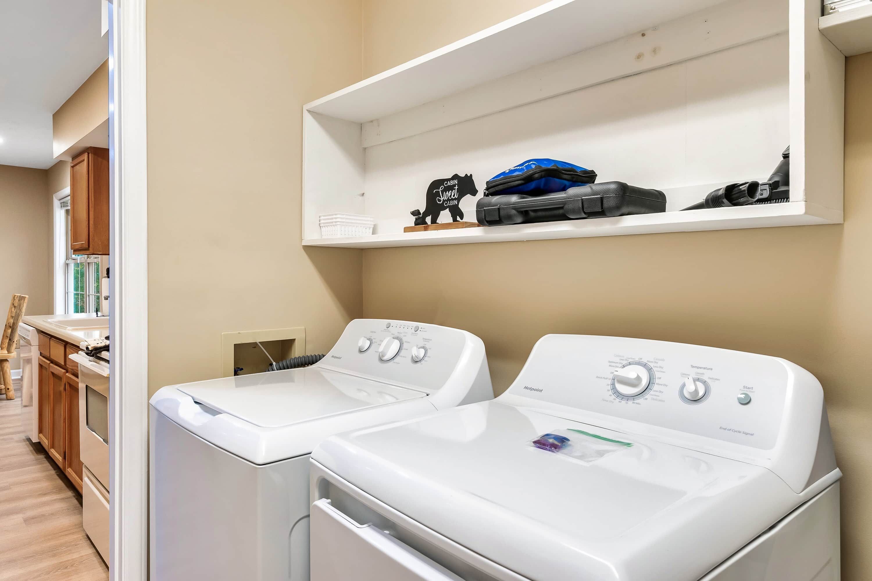 A laundry area with two front-loading washers and dryers, a shelf with a suitcase and a teddy bear, and wooden cabinets in the background.