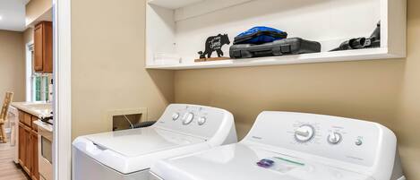 A laundry area with two front-loading washers and dryers, a shelf with a suitcase and a teddy bear, and wooden cabinets in the background.
