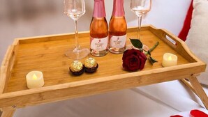 Un vassoio di legno con due bicchieri, due bottiglie, una candela, due cioccolatini confezionati, una rosa e un palloncino a forma di cuore su un letto.