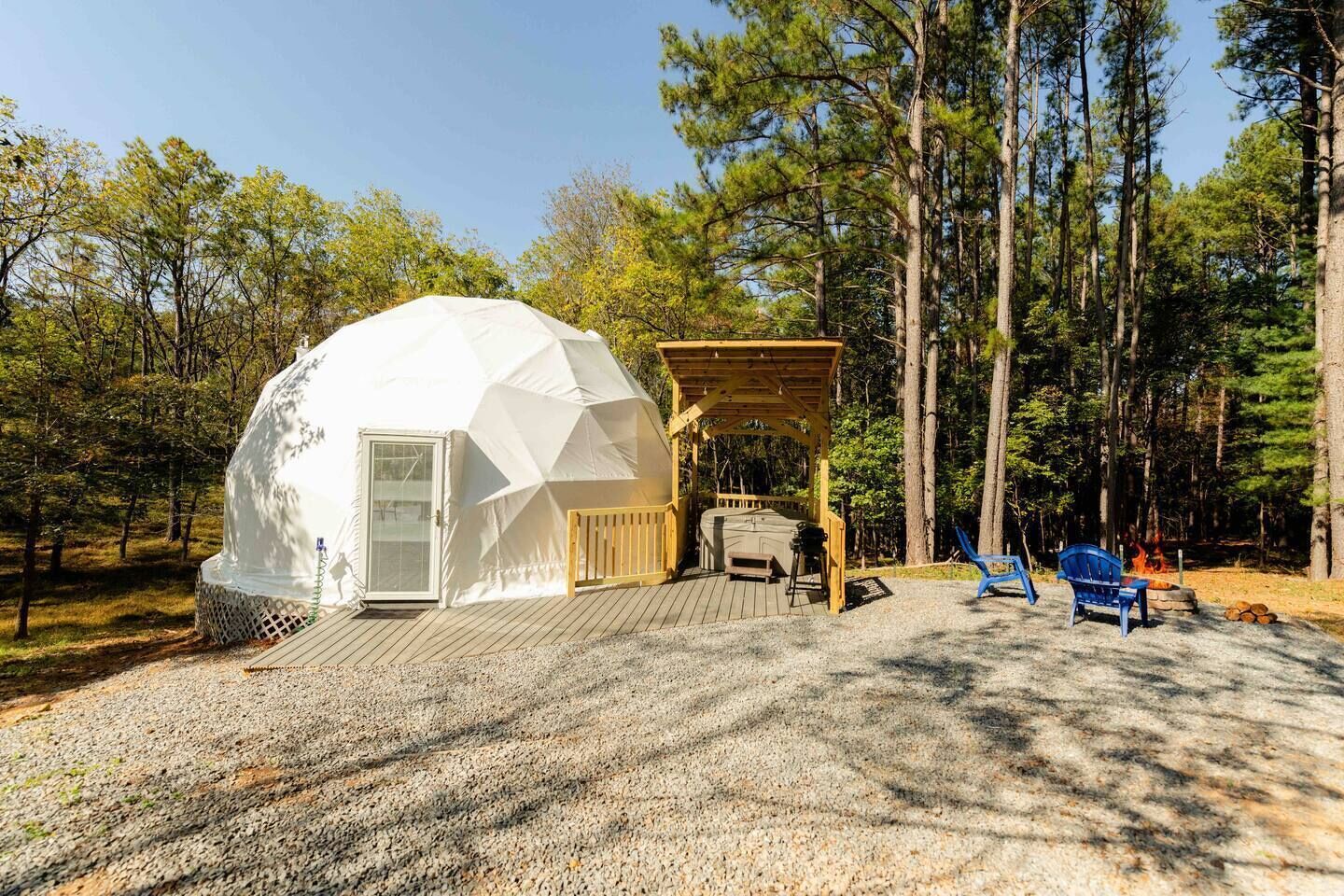 A geodesic dome structure with a wooden deck, a hot tub, and two blue chairs in a forest setting.