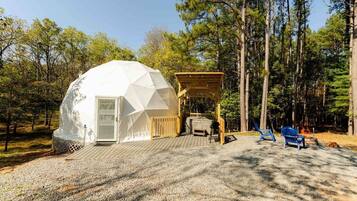 A geodesic dome structure with a wooden deck, a hot tub, and two blue chairs in a forest setting.