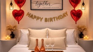 Una camera da letto con un letto, cuscini, un vassoio con bottiglie e bicchieri, palloncini a forma di cuore e uno striscione con scritto 'Happy Birthday'.