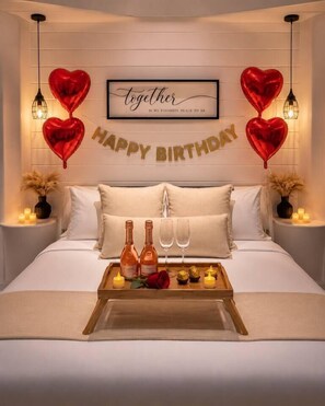 A bedroom with a bed, pillows, a tray with bottles and glasses, heart-shaped balloons, and a 'Happy Birthday' banner.