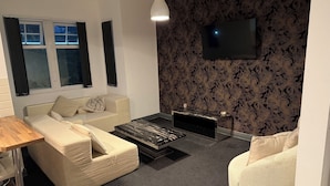 A modern living room with a sofa, a coffee table, a TV mounted on the wall, and a window with curtains. - Spacious Wakefield Getaway 3br (Wrenthorpe)