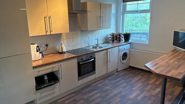 A modern kitchen with white cabinets, a built-in oven, a washing machine, and a window with a view of greenery. - Spacious Wakefield Getaway 3br (Wrenthorpe)