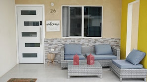A room with a white door, a welcome sign, a number 26, a wicker sofa with cushions, and a glass coffee table. - Casa de Paz
House of Peace (Little London)