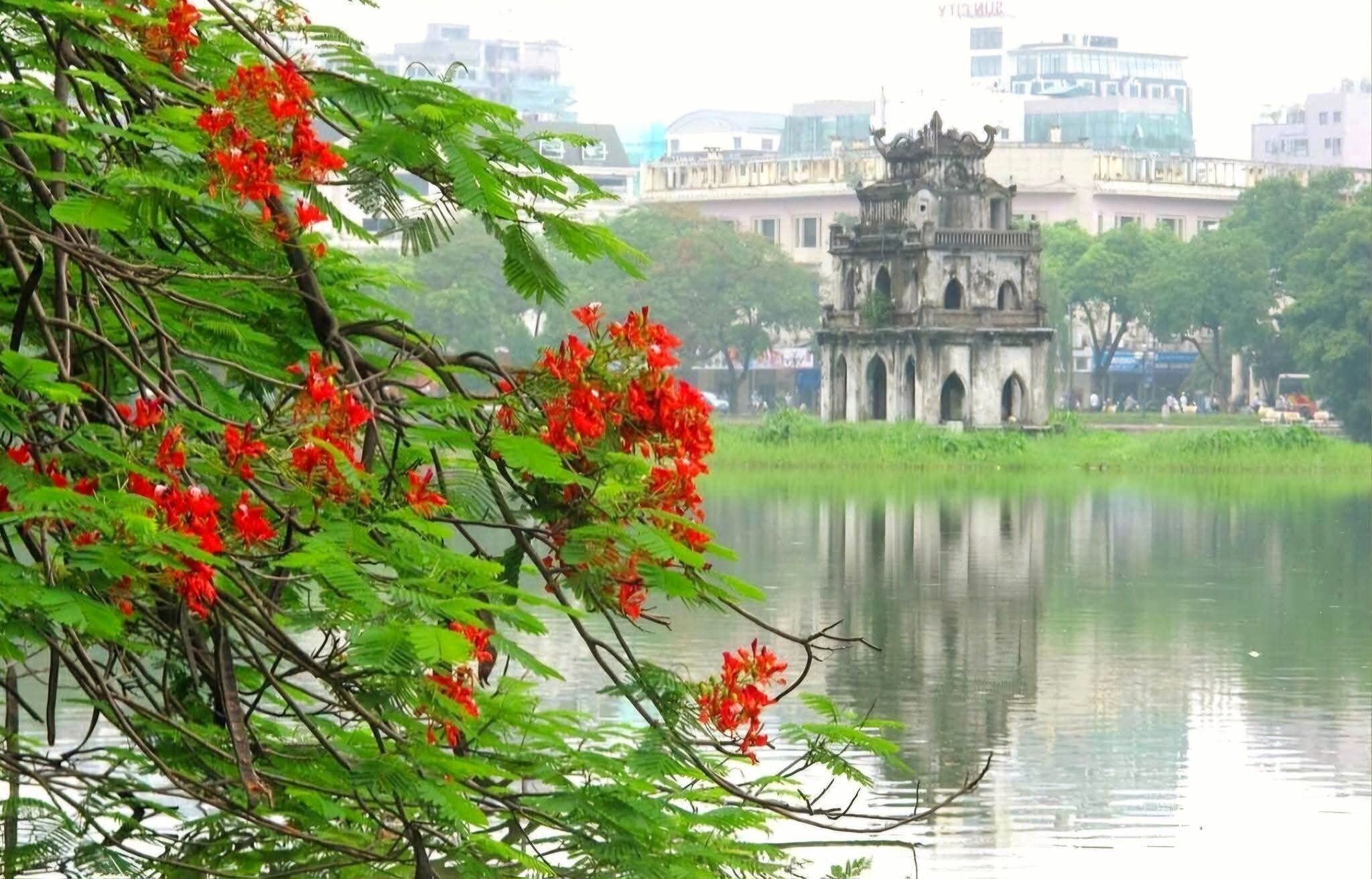 A traditional temple with a reflection in the water, surrounded by greenery and a cityscape in the background.