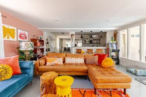 Ein modernes Wohnzimmer mit Sofa, Hocker und Couchtisch, im Hintergrund eine Küche und ein großes Fenster, das für natürliches Licht sorgt.