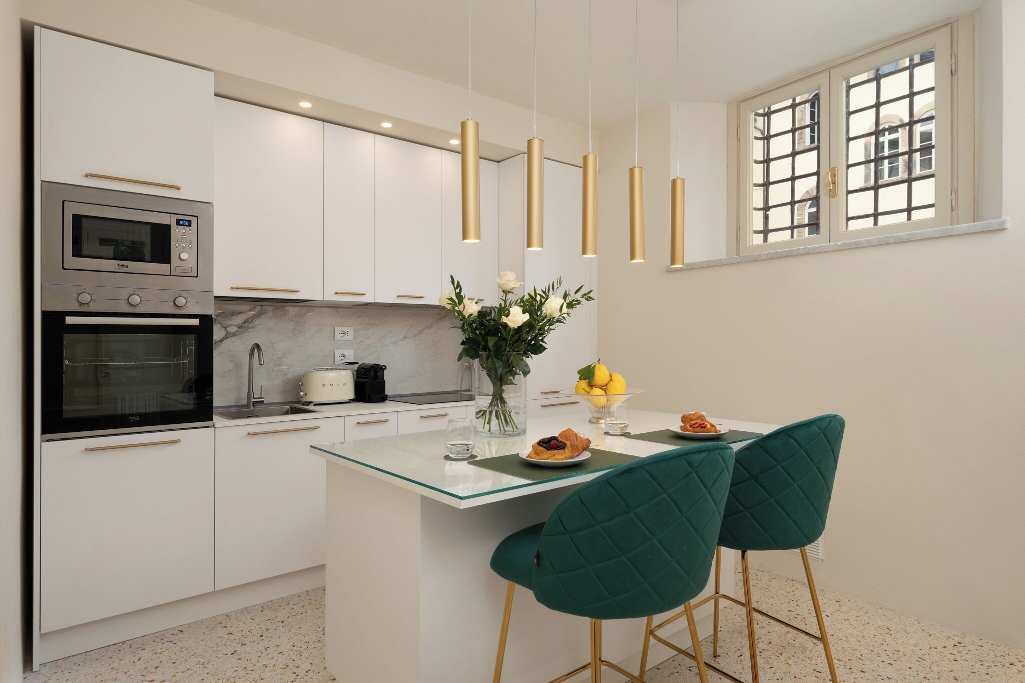 Eine moderne Küche mit weißen Schränken, einem eingebauten Backofen, einer Mikrowelle, einer Glasinsel und zwei grünen Stühlen.
