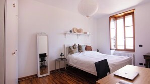 Ein Schlafzimmer mit einem Bett, einem Schreibtisch mit Laptop, einem Spiegel, einem Regal mit Dekoration und einem Fenster mit Tageslicht.