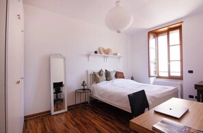 A bedroom with a bed, a desk with a laptop, a mirror, a shelf with decorative items, and a window with natural light. - Charming roof terrace apartment by the lake promenade (Salò)