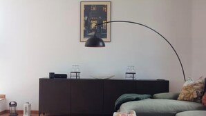 Ein modernes Wohnzimmer mit einem dunkelgrauen Sofa, einem Couchtisch mit Obstschale, einer schwarzen Stehlampe, einer Kommode mit Dekoration und einem gerahmten Bild an der Wand.