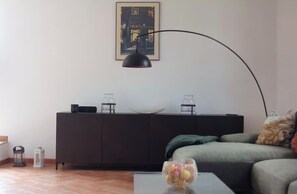A modern living room with a dark-colored sofa, a coffee table with a fruit bowl, a black floor lamp, a cabinet with decorative items, and a framed picture on the wall. - Charming roof terrace apartment by the lake promenade (Salò)