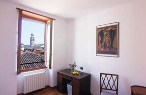 A room with a window overlooking a cityscape, a radiator, a desk with a vase, and a poster on the wall. - Charming roof terrace apartment by the lake promenade (Salò)