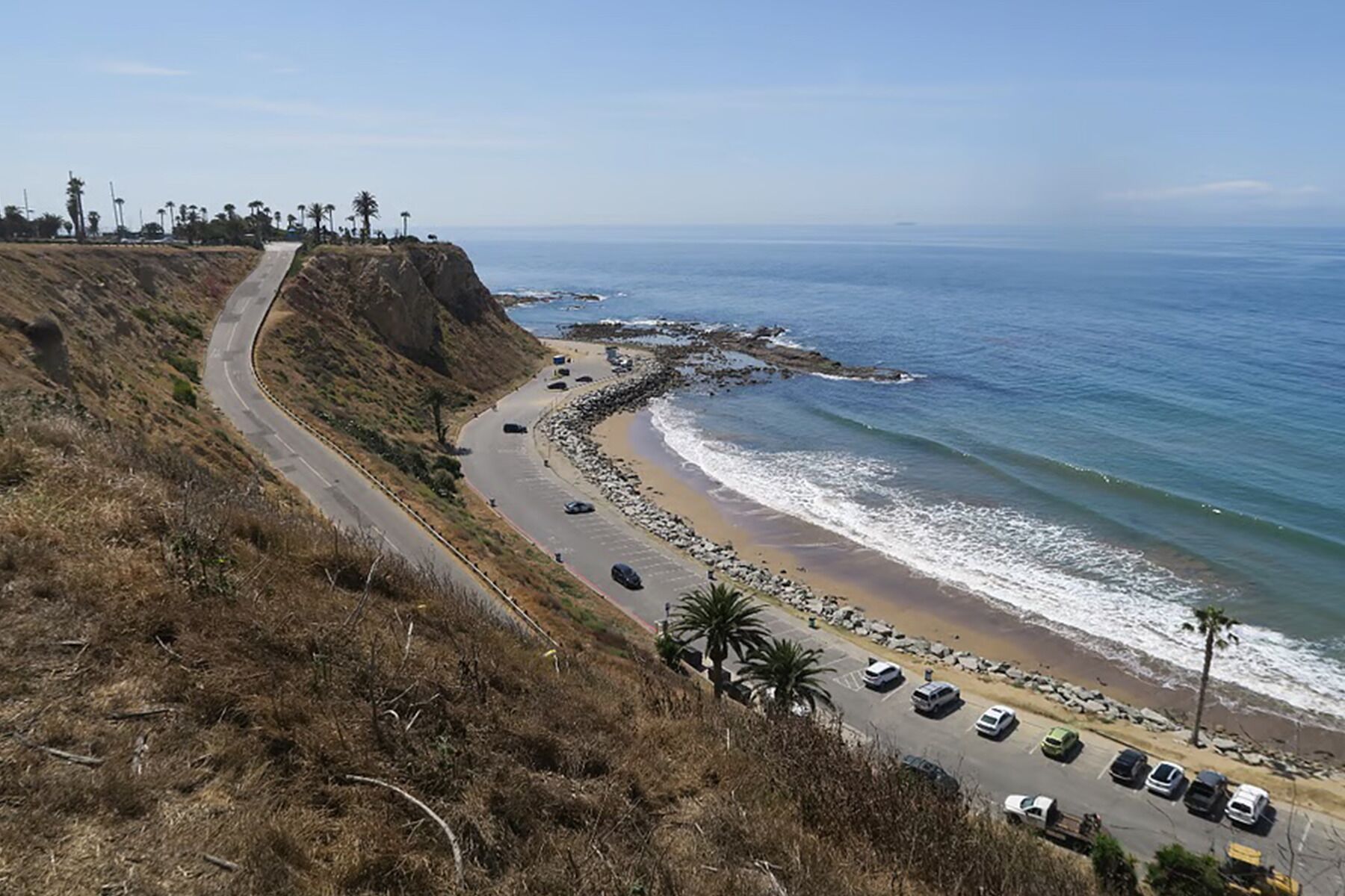 A coastal road with a beach, parked cars, and a clear sky.