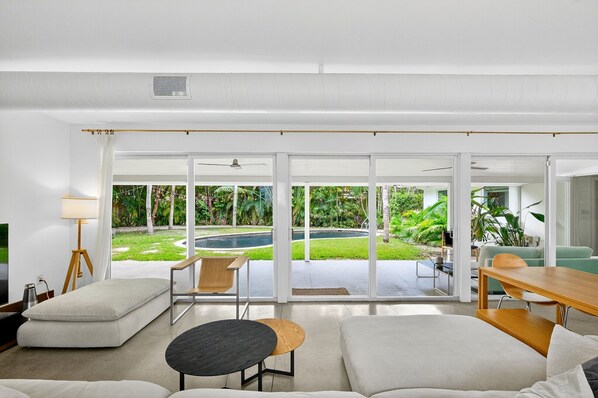 Ein modernes Wohnzimmer mit einer großen Schiebeglastür, die zu einem Poolbereich führt, einer Couch, einem Couchtisch und einer Lampe.