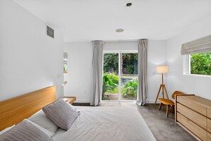 Ein Schlafzimmer mit Bett, Schreibtisch, Lampe und Blick auf Grünfläche.