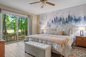 Une chambre avec un grand lit, deux lampes de chevet, un ventilateur de plafond et une vue sur un jardin, accessible par une porte-fenêtre coulissante.