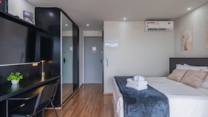 A modern hotel room with a bed, bedside table, a desk with a chair, an air conditioning unit, and a small plant. - Excellent Studio in S o Paulo - Sp-a1107 (São Paulo)