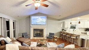 Una sala de estar con chimenea, televisor, ventilador de techo y una zona de cocina con taburetes de bar.