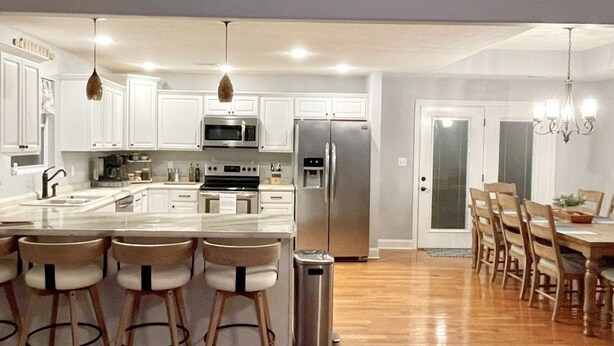 Una cocina moderna con armarios blancos, electrodomésticos de acero inoxidable, una isla central con taburetes y un comedor con sillas de madera.