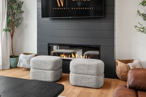 Un salon moderne avec une cheminée, des poufs gris à revêtement en tissu, un téléviseur à écran plat et une plante en pot.