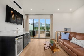 Un salon moderne comprenant une cuisine, un canapé en cuir marron, une table basse en bois et une grande porte-fenêtre coulissante donnant sur un balcon extérieur.