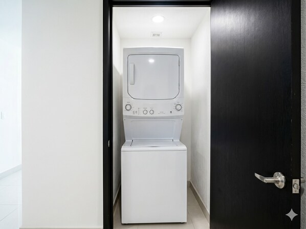 Máquina de lavar compacta e branca, fixada na parede, em um corredor estreito com uma porta escura à direita.