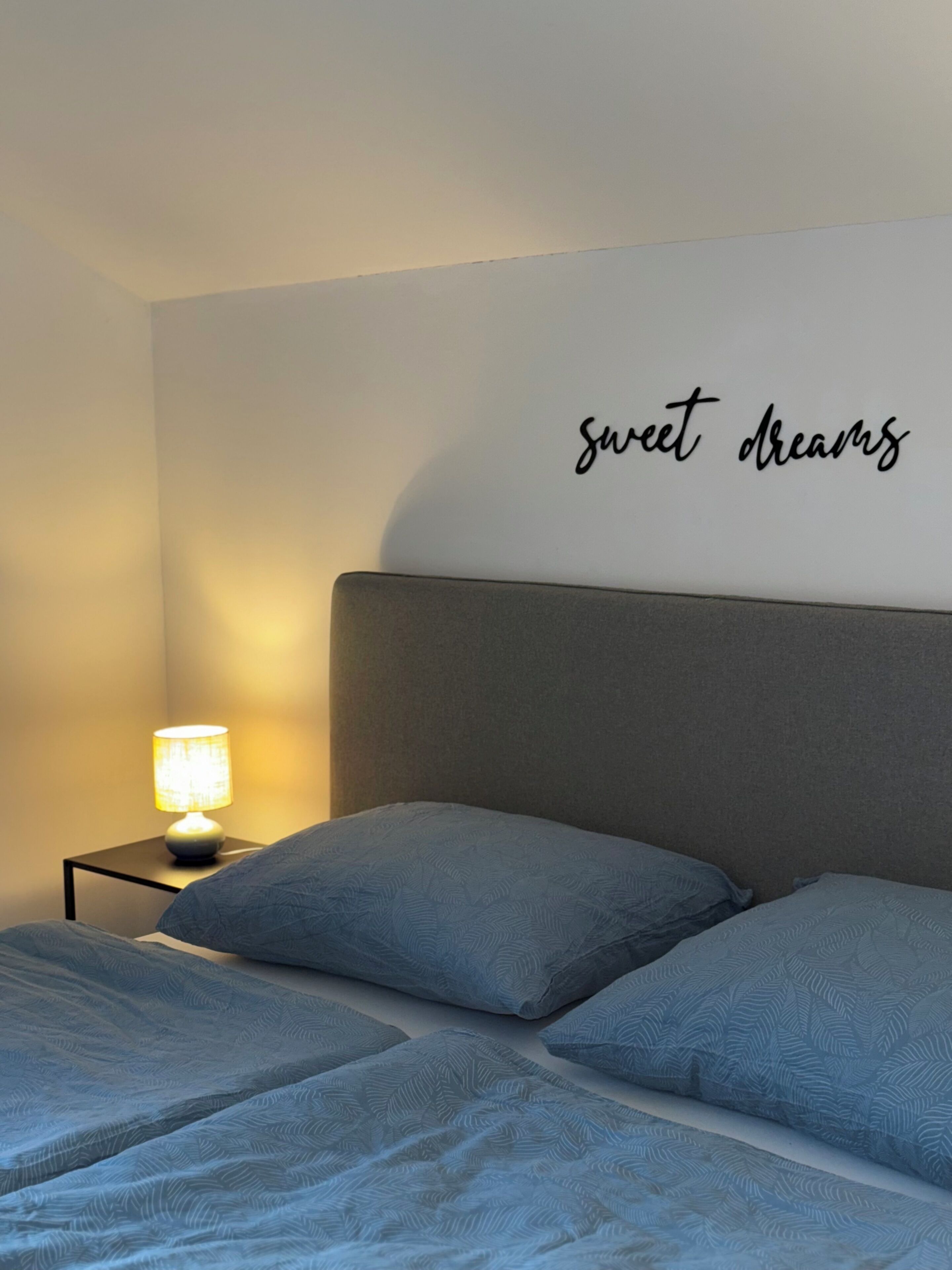 A bedroom with a bed, pillows, a nightstand with a lamp, and a wall with the text 'sweet dreams'.
