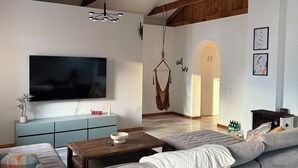 Een moderne woonkamer met een grote grijze bank, een houten plafond, een flatscreen televisie, een hangstoel en een blauwe kast.