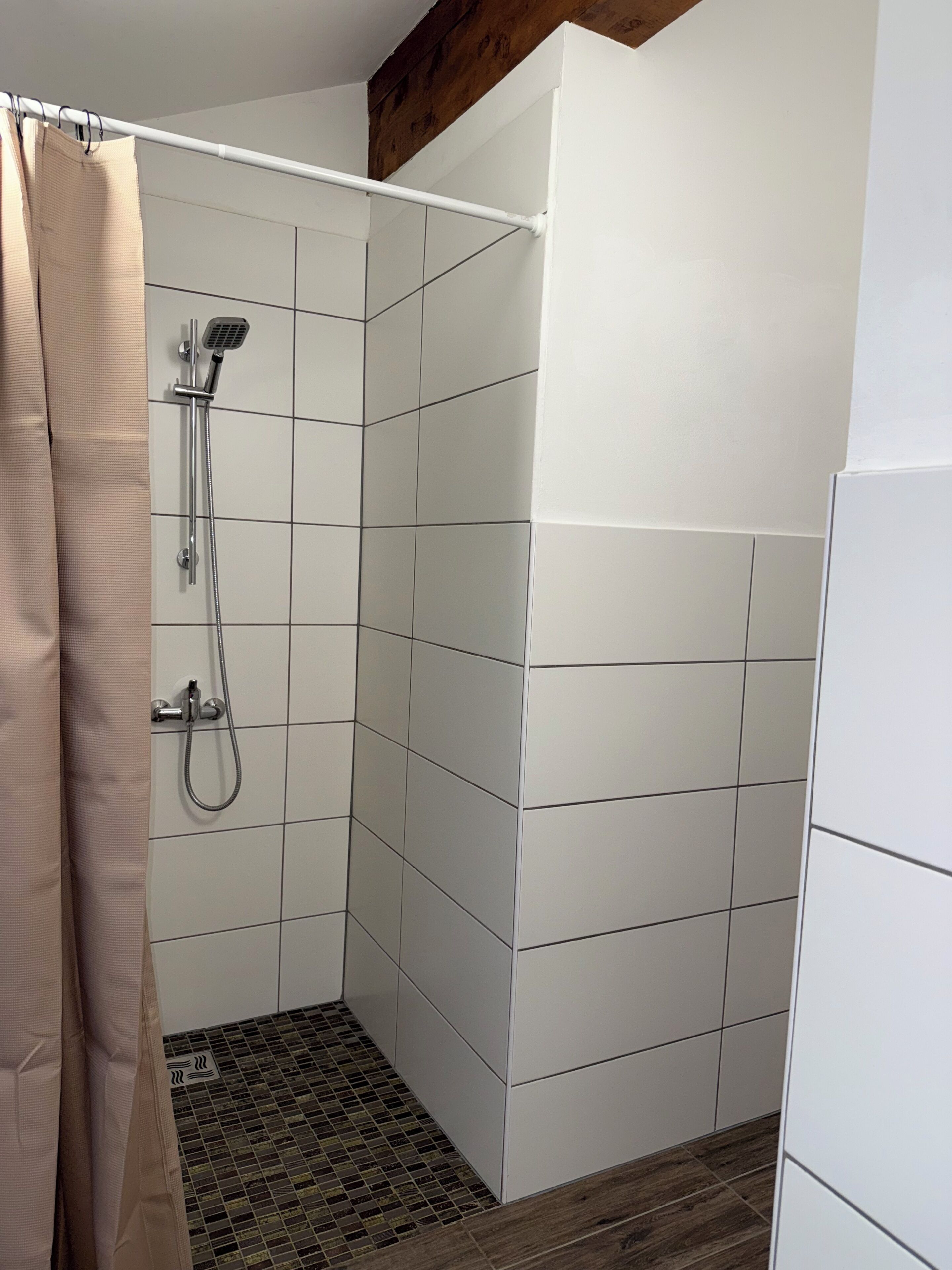 A shower with a handheld showerhead and a wall-mounted faucet, tiled walls, and a wooden beam ceiling.