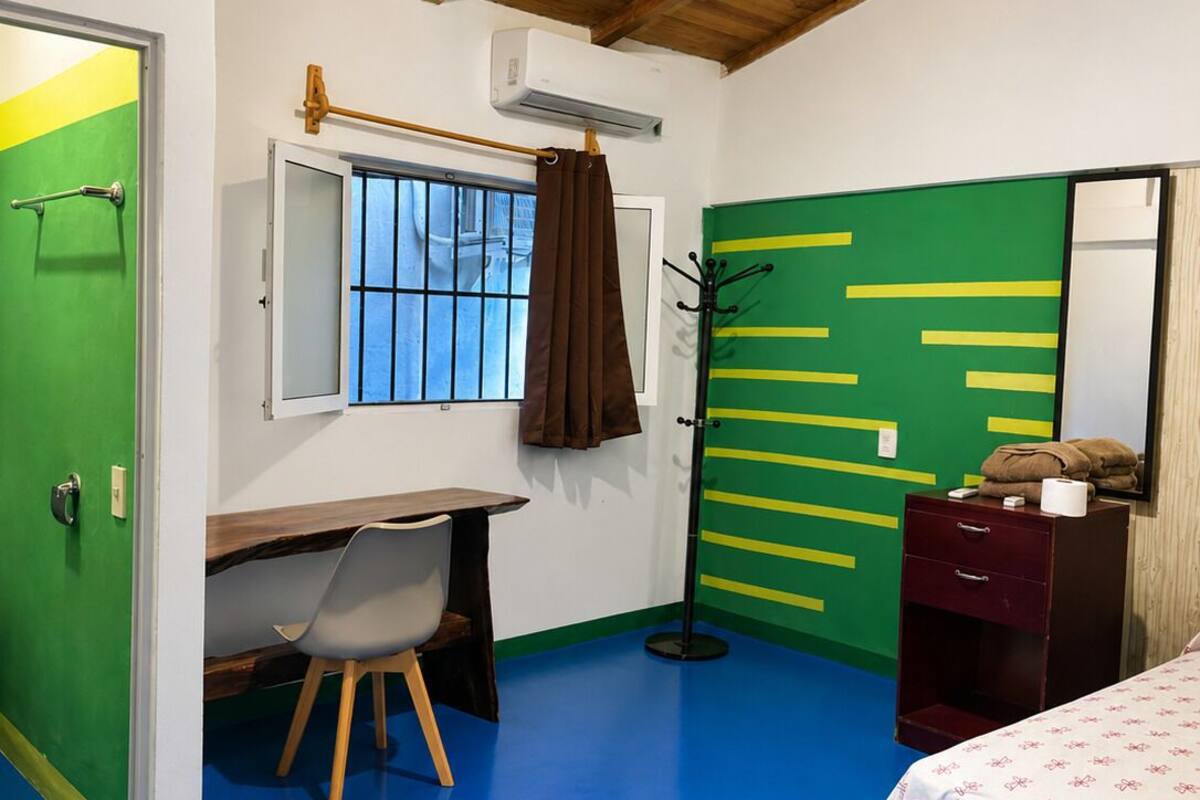 A room with a wooden ceiling, a desk with a chair, a bed, a dresser, and a window with a curtain.