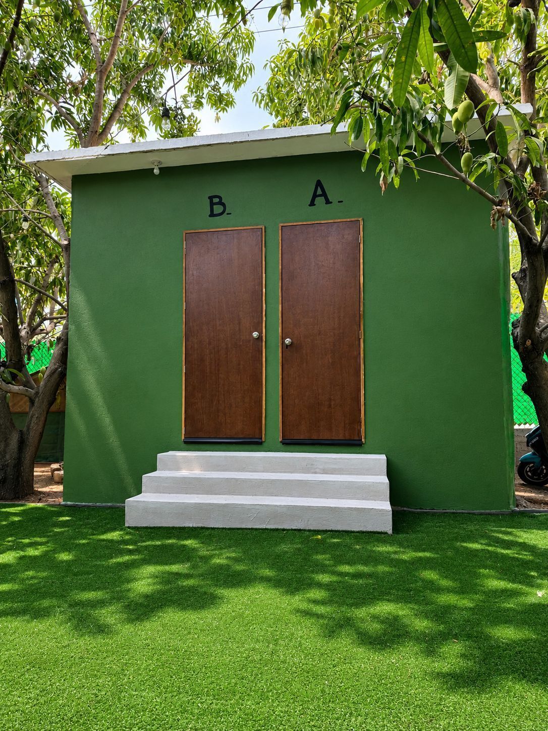 Un pequeño edificio verde con dos puertas de madera identificadas con las letras 'B' y 'A', y un conjunto de escalones blancos que conducen a ellas.