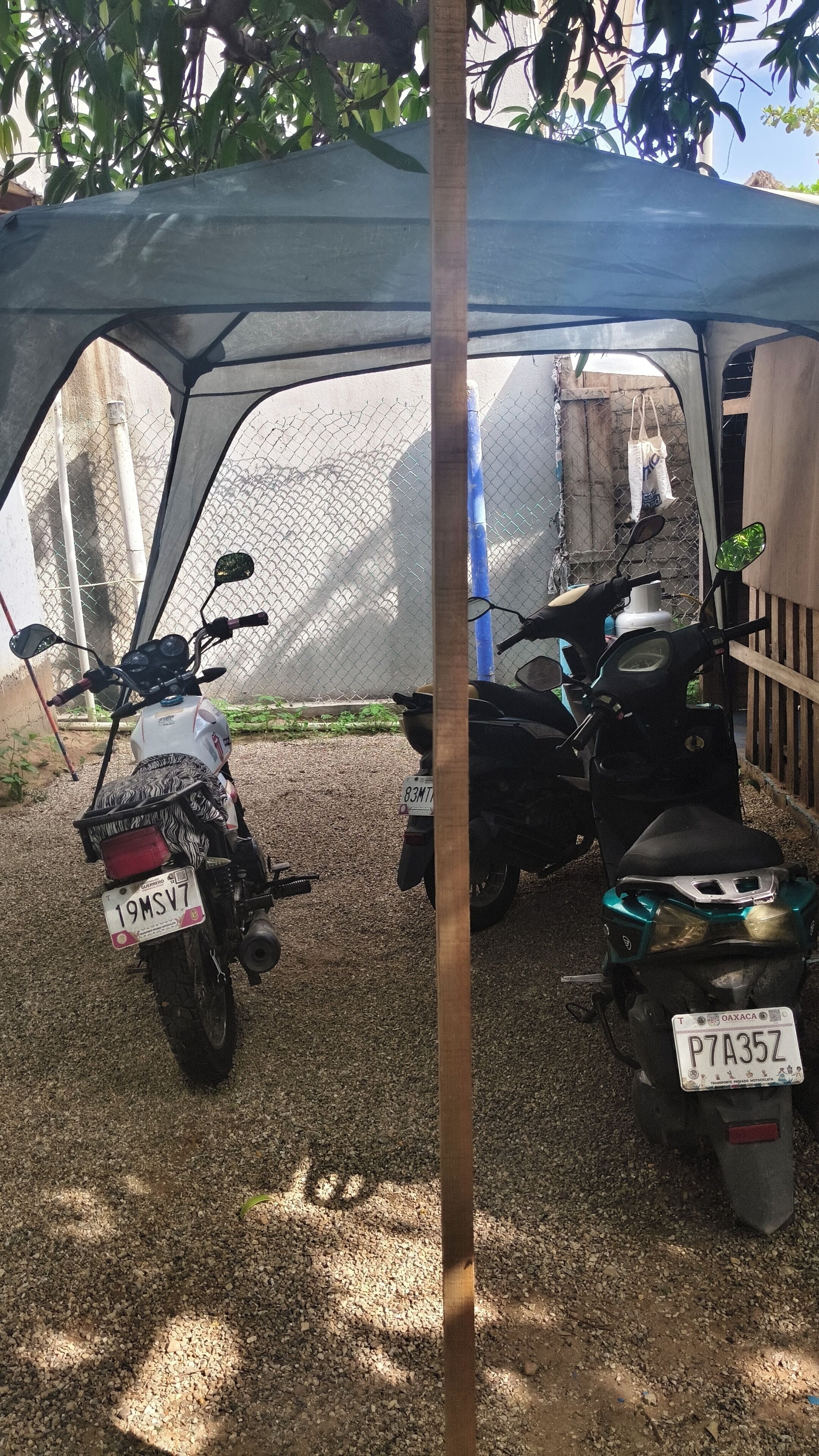 Dos motocicletas estacionadas bajo un techo, con una cerca de malla metálica al fondo.