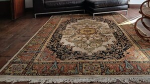 Uma sala de estar com sofá de couro preto, tapete estampado, lustre e quadros nas paredes.