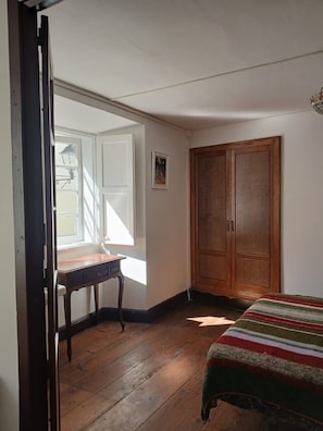 Ein Zimmer mit einem Holzschrank, einem Bett mit gestreifter Decke, einem Schreibtisch und einem Fenster mit Jalousien.