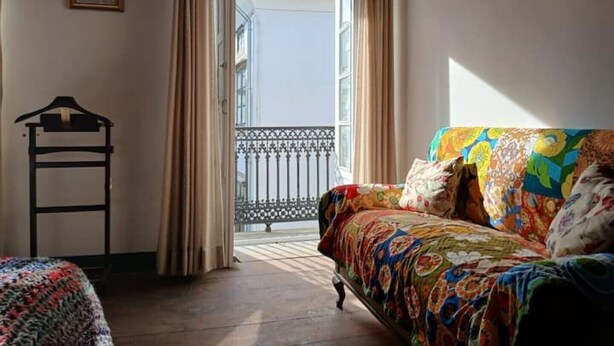 Quarto com cama, sofá colorido, cadeira de madeira e varanda com grade de metal.