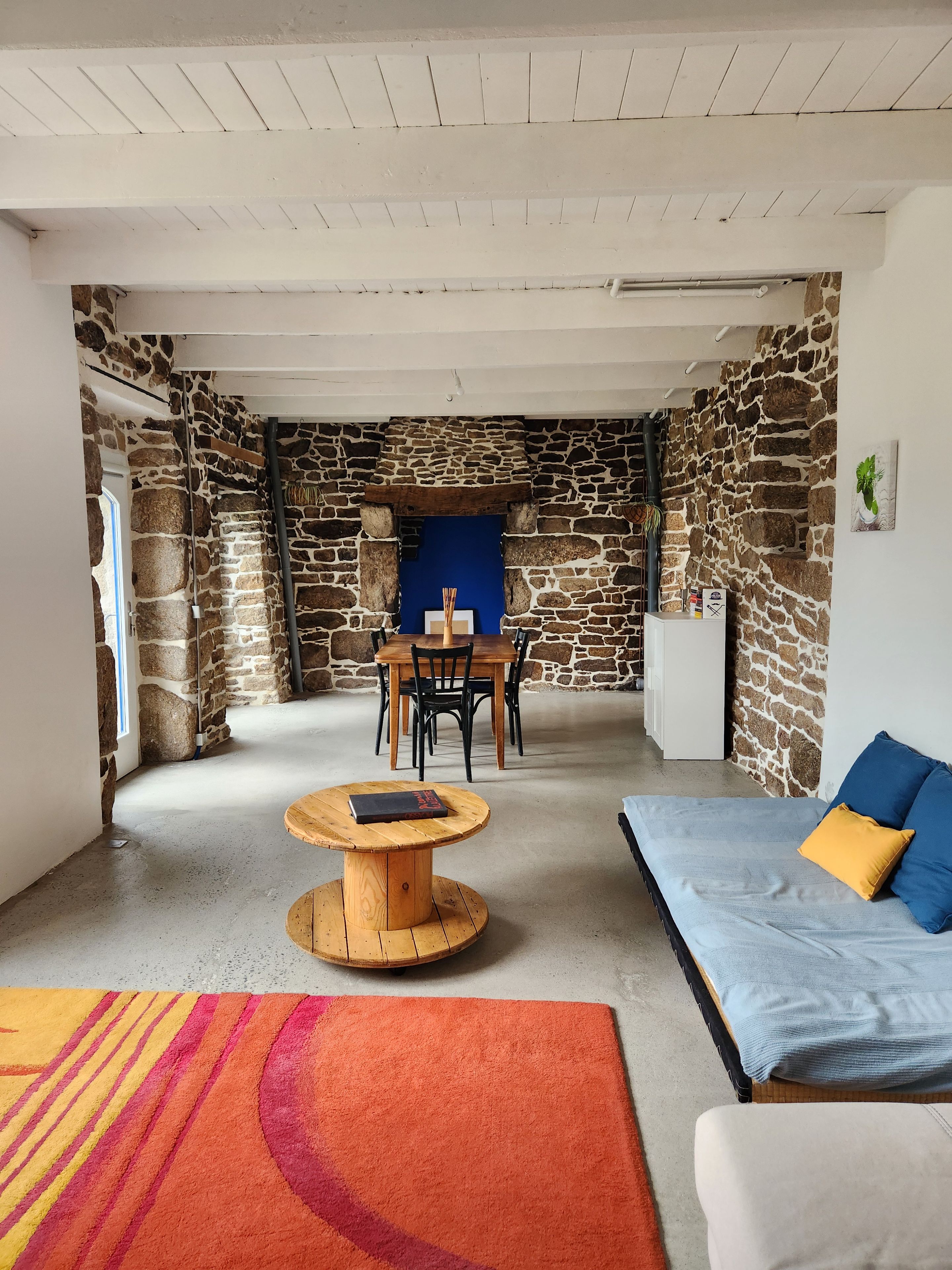 Een kamer met een stenen muur, een eettafel met stoelen, een ronde houten tafel, een bank met kussens en een kleurrijk tapijt.