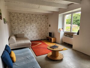 Een woonkamer met een bank, een televisie op een houten standaard, een ronde houten salontafel en een raam met uitzicht op groen.